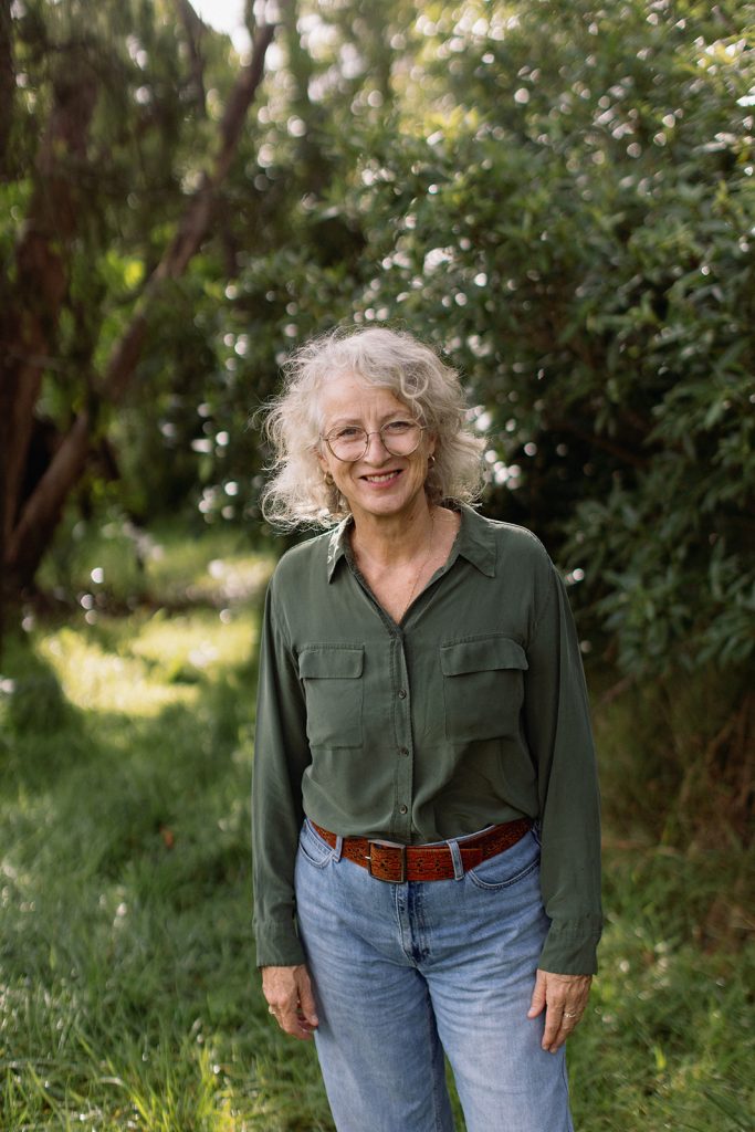 Portrait of Sue Lansdown standing in a sunlit grassy clearing, hands resting at her sides.